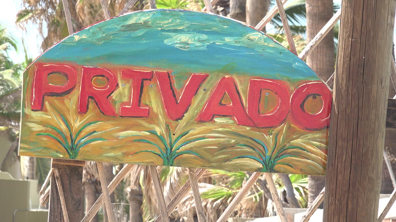 At 0:00, a close view of a hand-painted PRIVADO sign in Grand Junction, Colorado, shows red letters on a teal and yellow board, a wooden post on the right, palm trunks behind it, and no visible people.