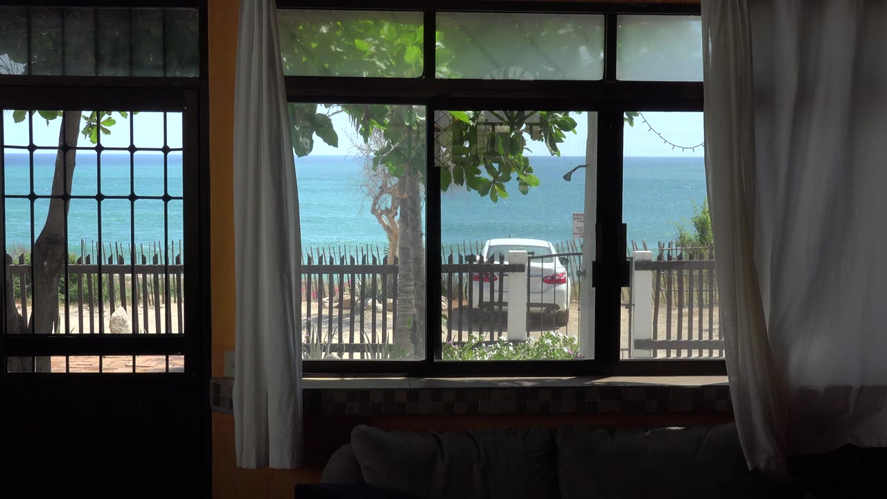 At 0:00, a shadowed house interior in Grand Junction Colorado frames a bright ocean view through barred windows and white curtains, with trees, a fence, a parked white car, and no visible people.