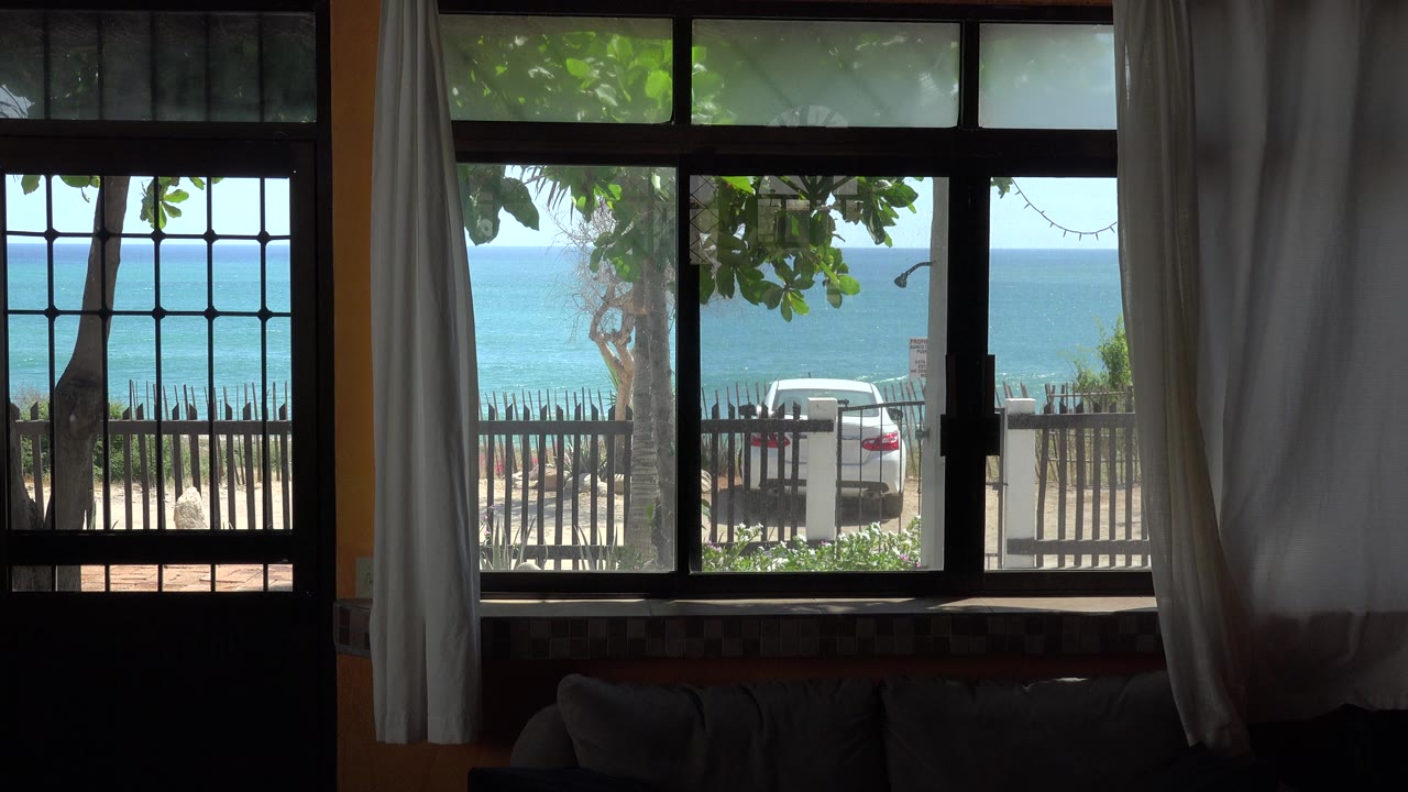 Dark room looking through a barred window toward trees, a fence, a white car, and blue water.