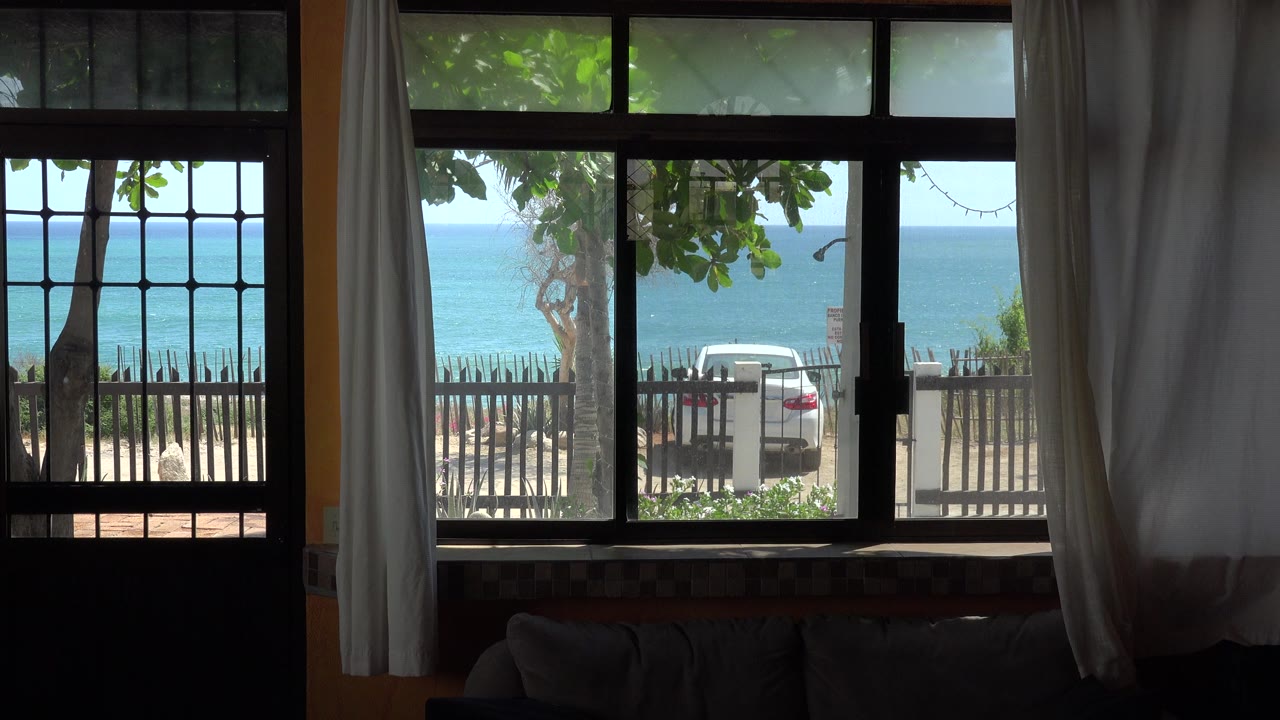 At 0:01, the Grand Junction Colorado collection view remains framed by white curtains and black window bars, with a white car behind a fence, bright blue water beyond it, and no visible people.