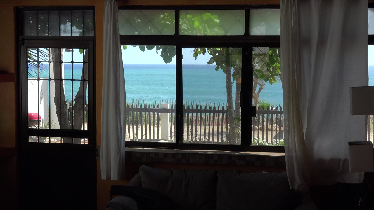 At 0:00, a shaded Grand Junction Colorado house interior frames a bright ocean view through a window and glass door, with curtains, trees, a fence, sand, sky, and no visible people.