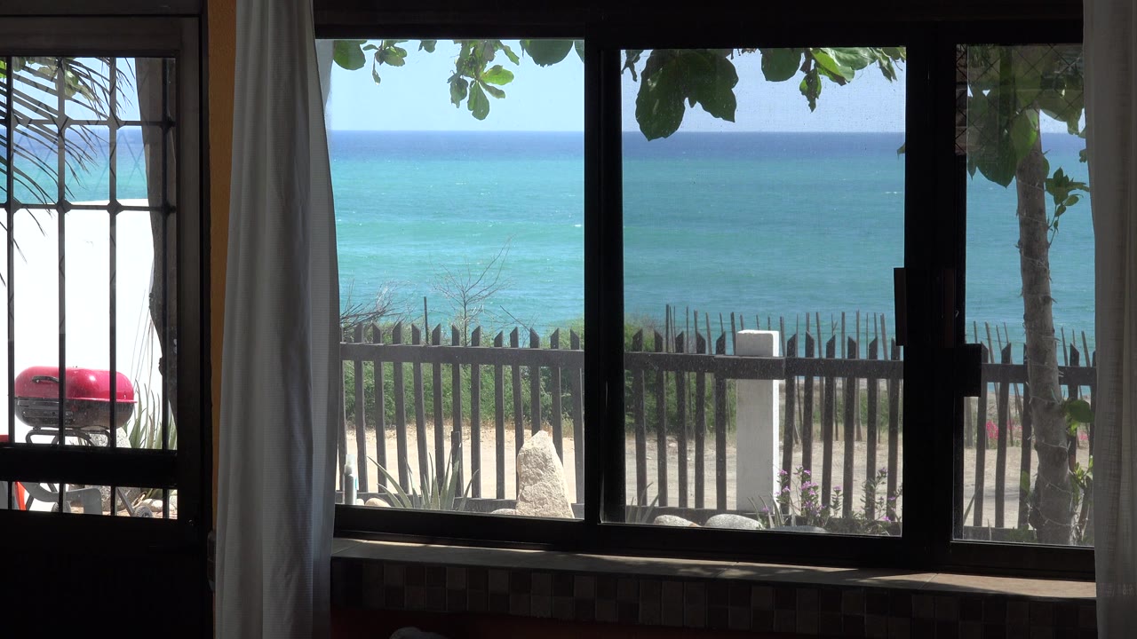 Interior window framing ocean, beach, fence, curtains, and a red outdoor grill-like object.
