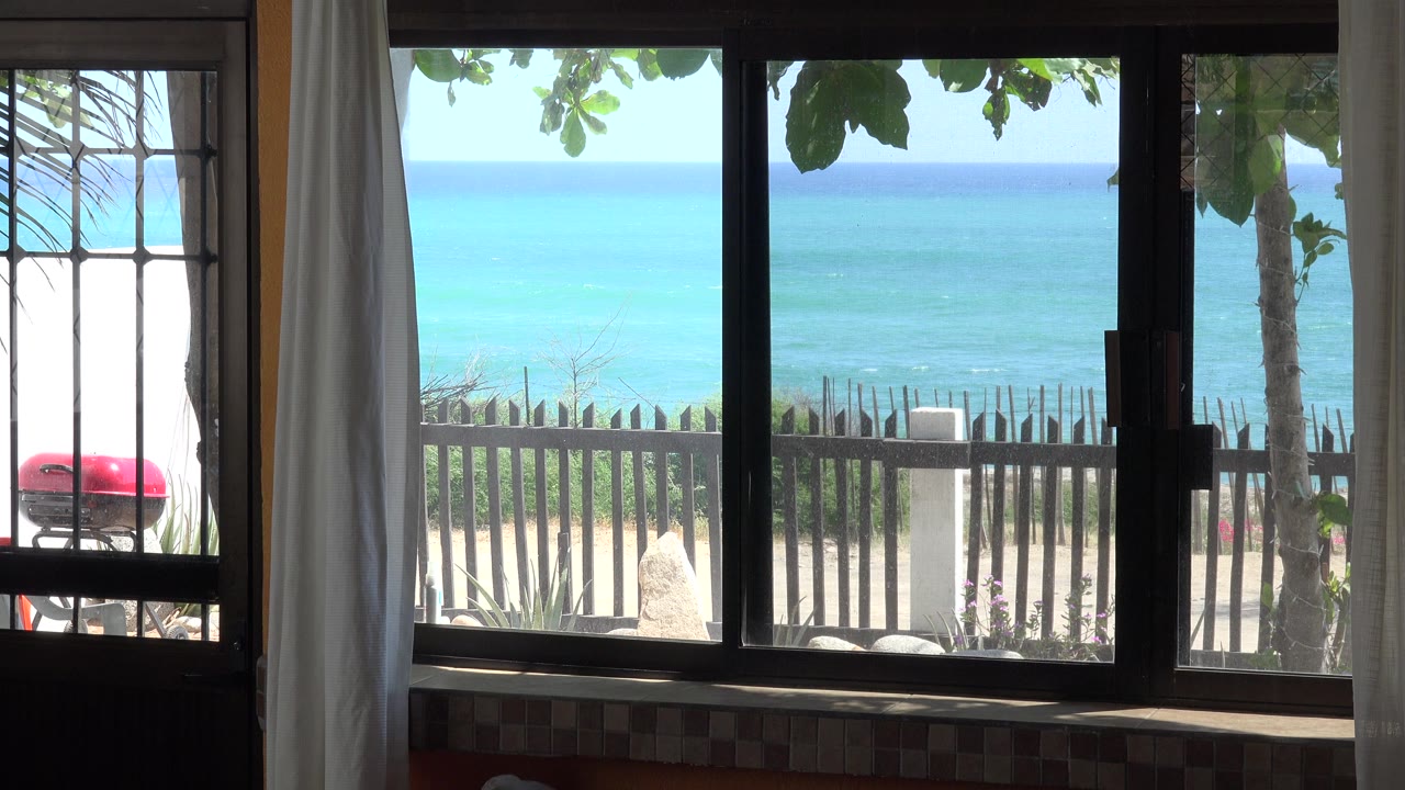 At 0:00, a shaded house interior in Grand Junction Colorado frames a bright beach and blue ocean through a window and side door, with no visible people.