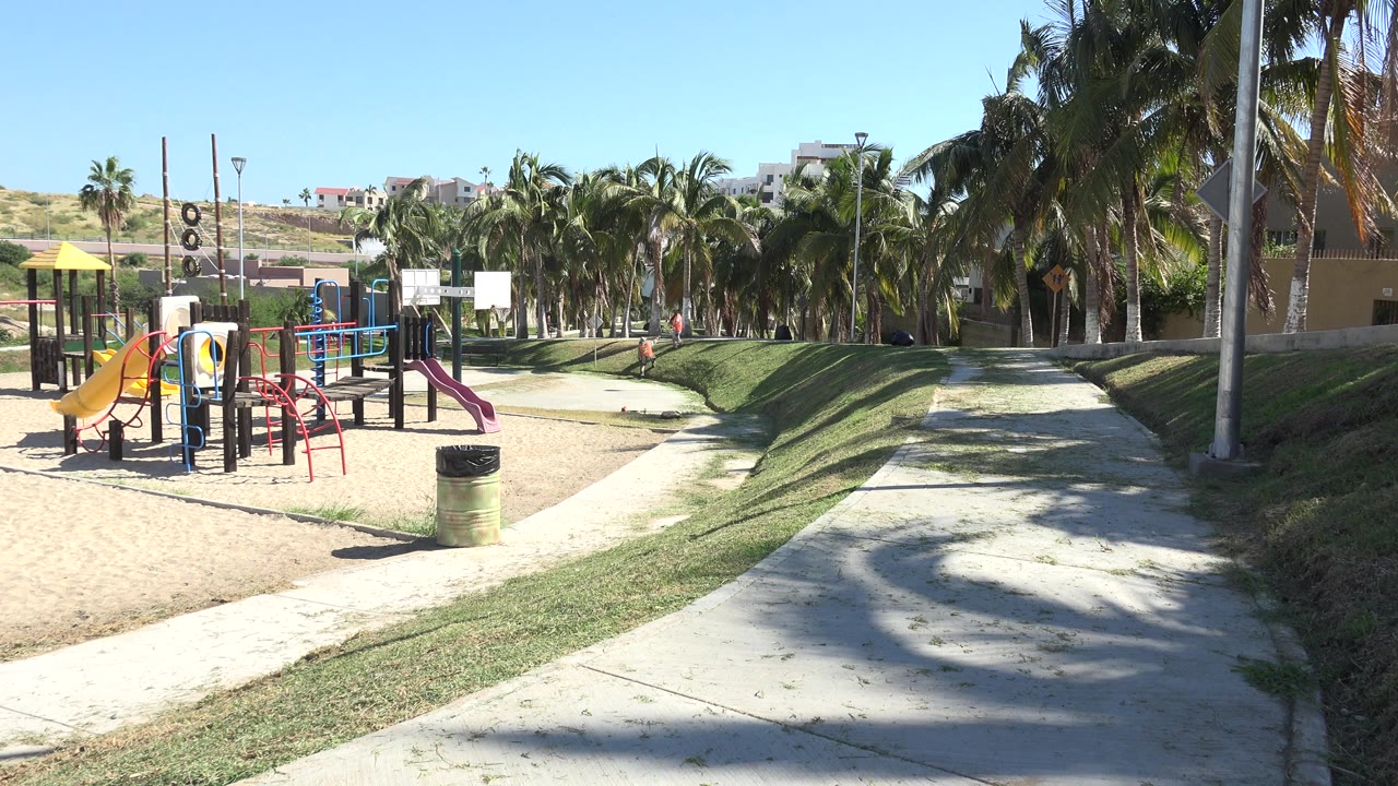 Sunny Grand Junction playground at the start of the clip, with yellow slides on sand, curving concrete paths, grass berms, palms, hillside buildings, and no visible people.