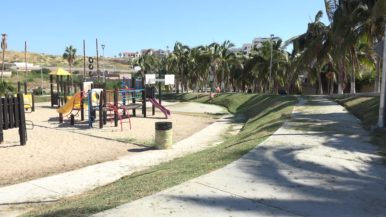 Playground with black climbing panel, slides, palms, and a child in the distance