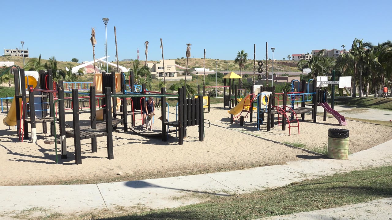 Wide sunny playground with slides, monkey bars, people, grass, and buildings beyond.