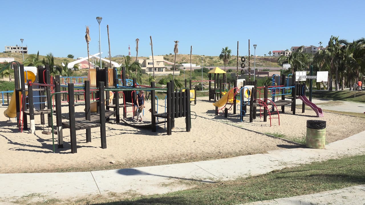 Grand Junction, Colorado park playground with adults and children visible near red climbing bars, yellow slides, black support posts, grass, palm trees, and buildings beyond the play area.