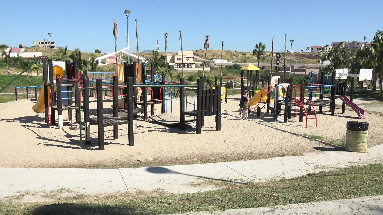A wide Grand Junction, Colorado playground view shows sand, slides, climbing structures, grass, sidewalks, palm trees, distant homes, and small people near the equipment.