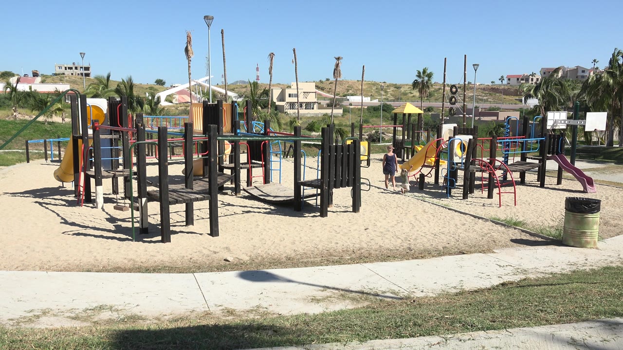 A broad view of the Grand Junction playground shows the same slides, platforms, rails, sand, and distant residential buildings, with people still visible near the central play equipment.