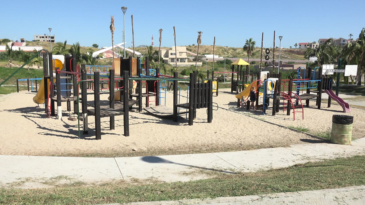 Sunny playground with slides, rails, sand, grass, and children near the equipment.