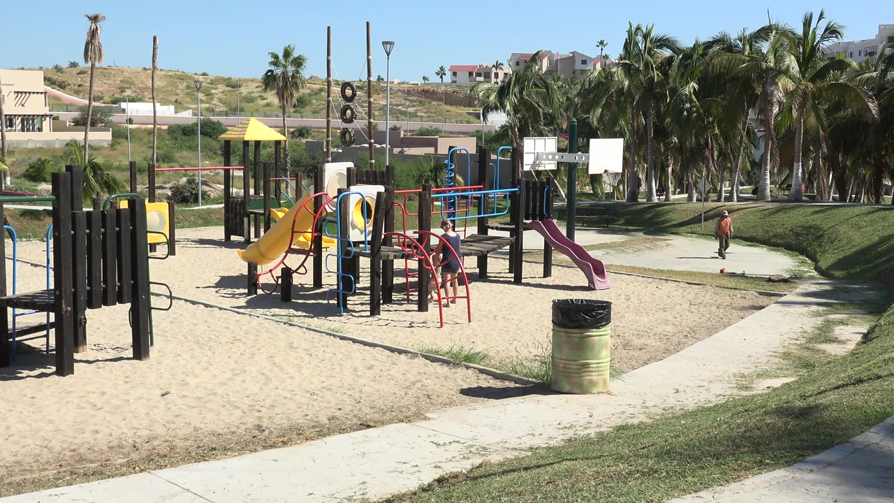 Wide playground with a child on a path near grass and palm trees.