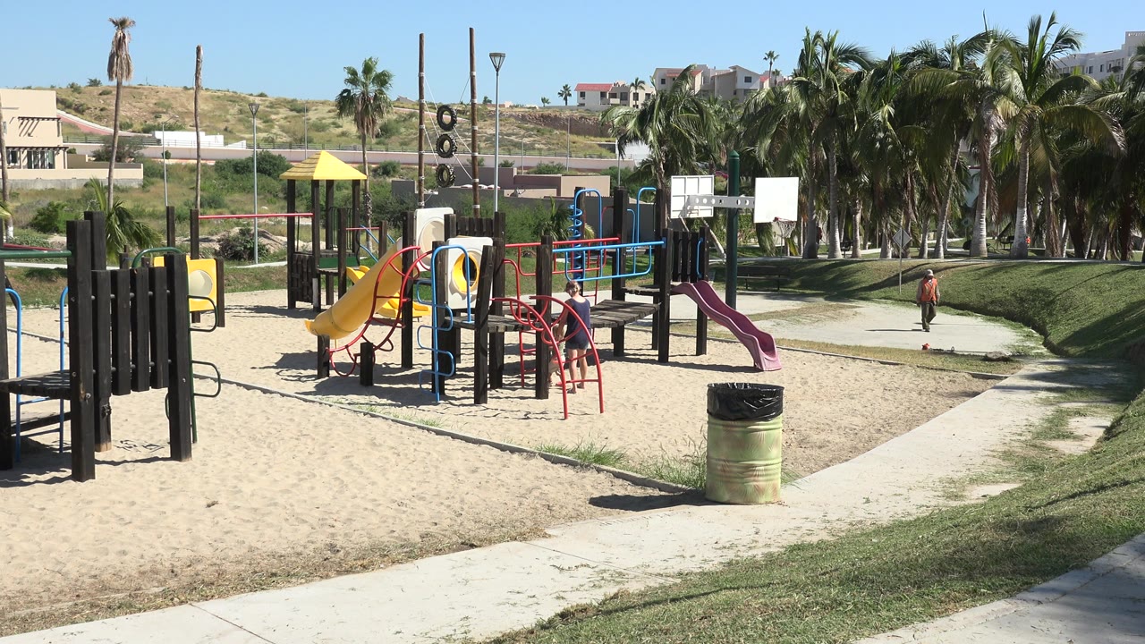 Grand Junction playground scene with the child slightly farther along the path, colorful play structures in sand, palms behind, and hillside homes across the background.