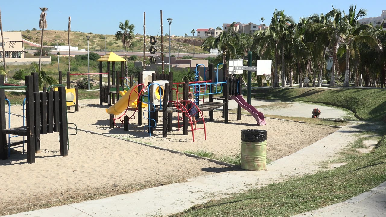 Sunny playground with slides, sand, palm trees, sidewalk, grass, and distant hills.