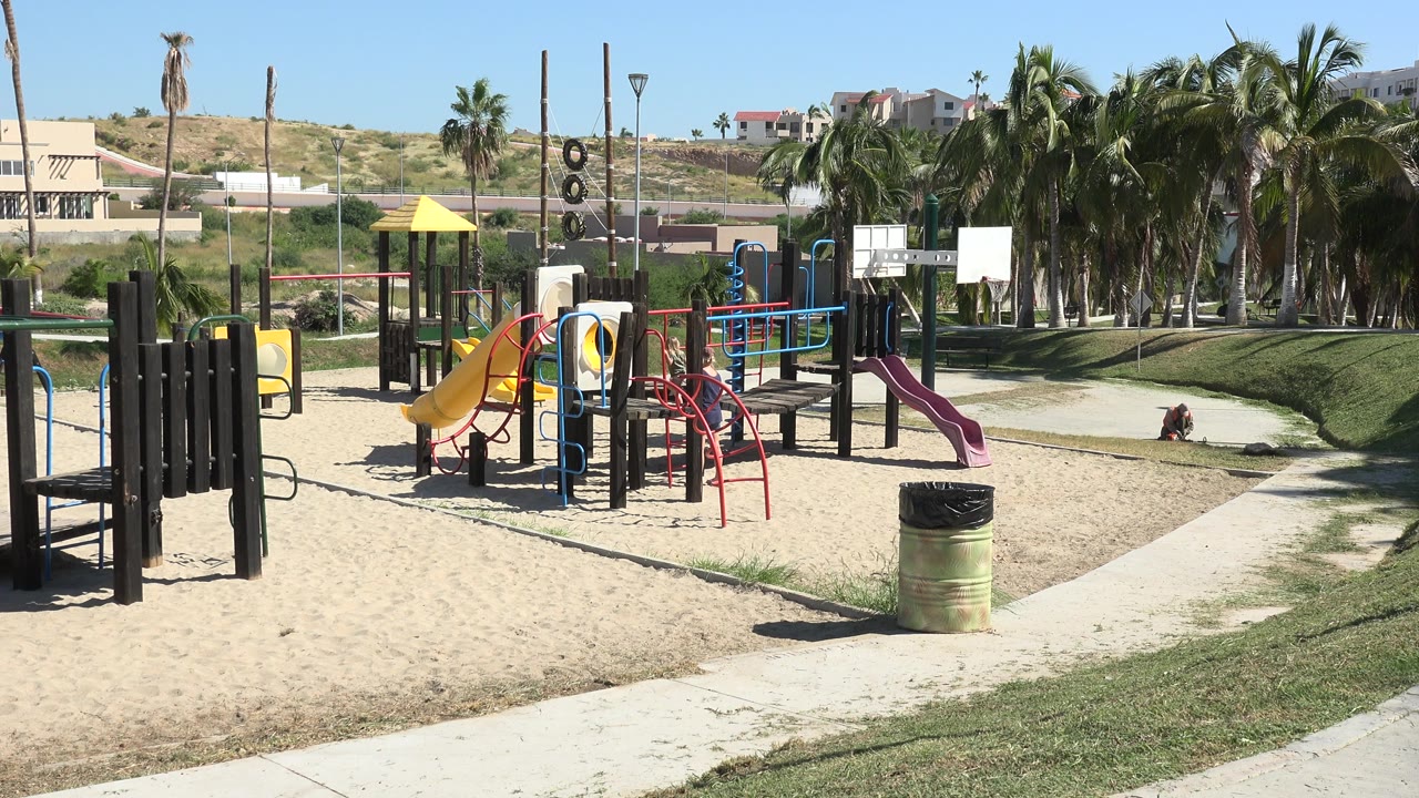 Grand Junction playground under bright sun with yellow and purple slides, red climbing bars, sand, a curving path, palm trees, distant hillside homes, and no visible people.