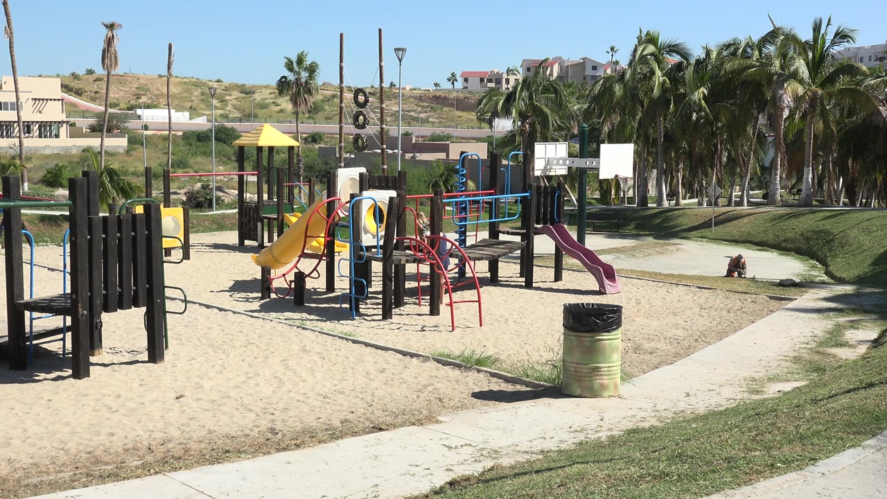Empty playground with colorful slides, palms, grass, and hillside homes.