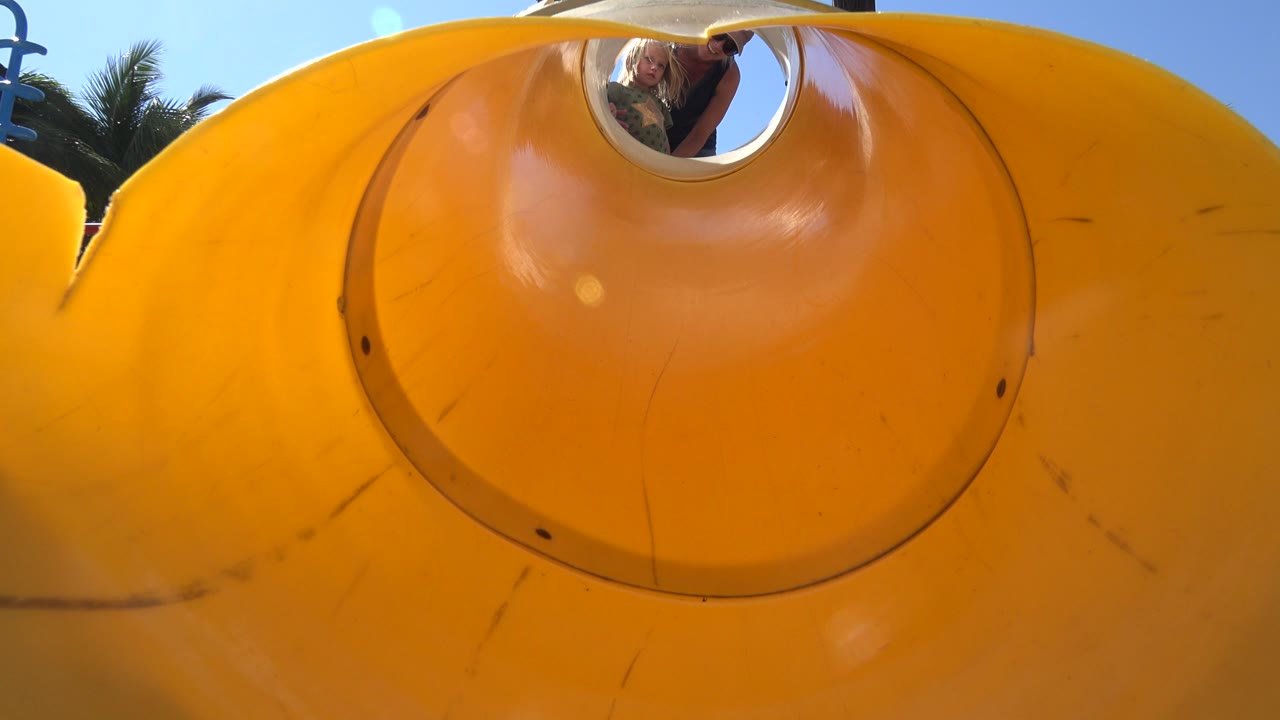 Ground-level view up a bright yellow playground slide in Grand Junction, Colorado, with an adult and child partly visible through the round top opening.