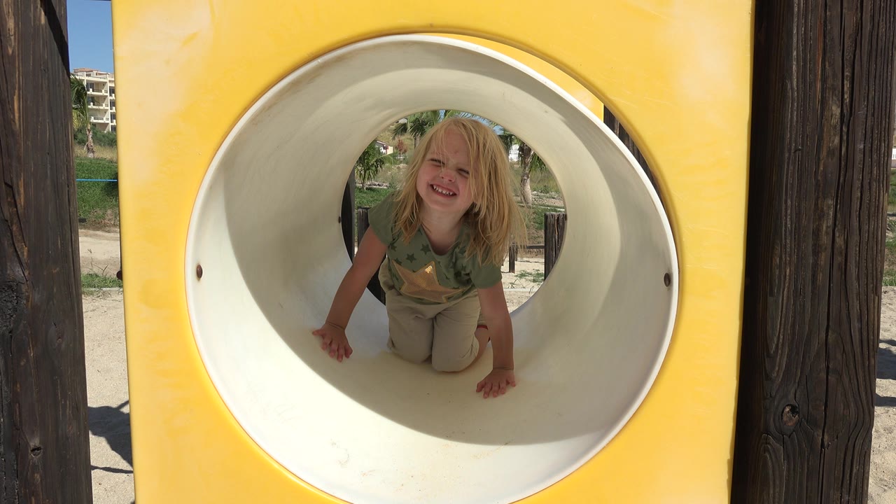 At 0:01, the child remains crouched inside the circular white slide opening, framed by yellow playground plastic and wooden posts.