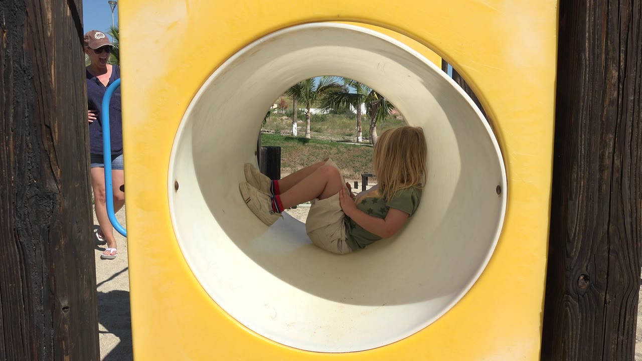 At 0:01, the child remains seated inside the circular tunnel while bright sun hits the yellow playground panel and trees are visible through the far opening.