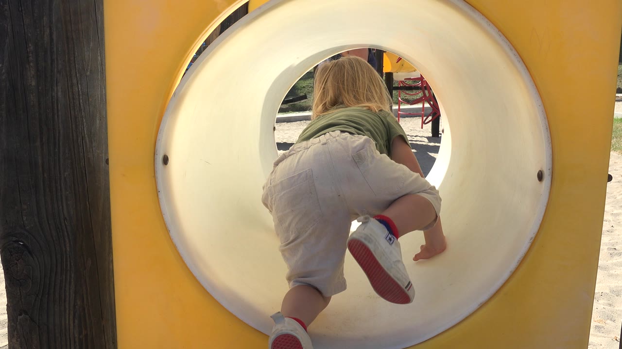 A blonde child in a green shirt and light shorts crawls away from the camera inside a white-rimmed yellow playground tunnel in Grand Junction, Colorado.