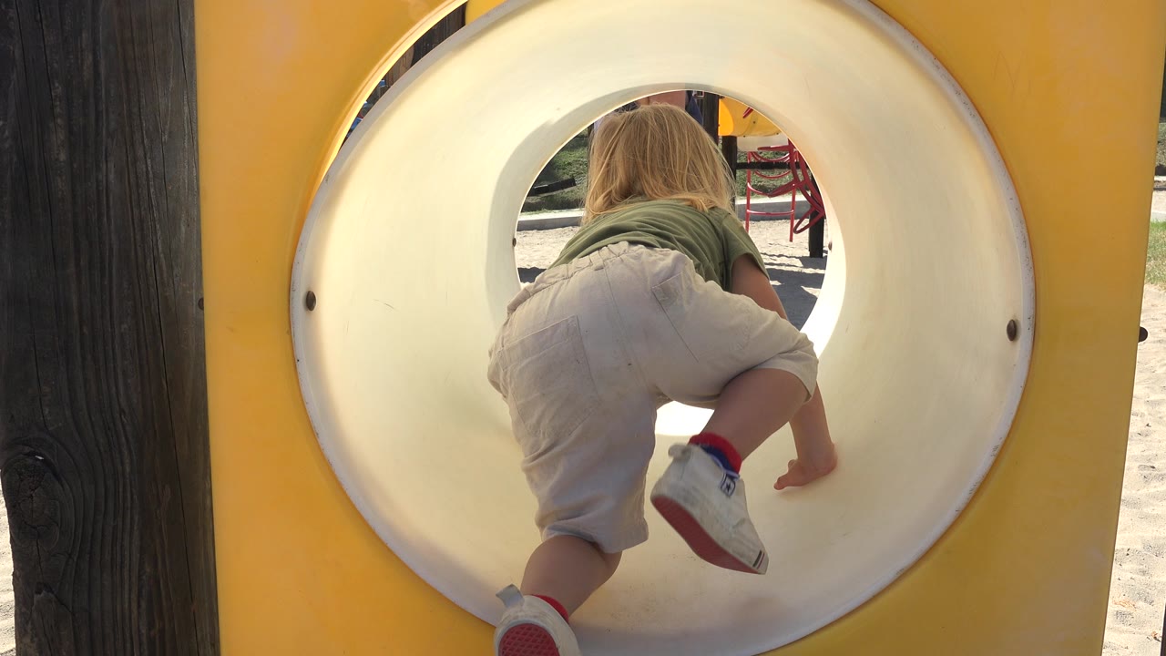 The blonde child continues crawling through the yellow playground tunnel, with one shoe lifted near the camera and sand visible beyond the opening in Grand Junction, Colorado.