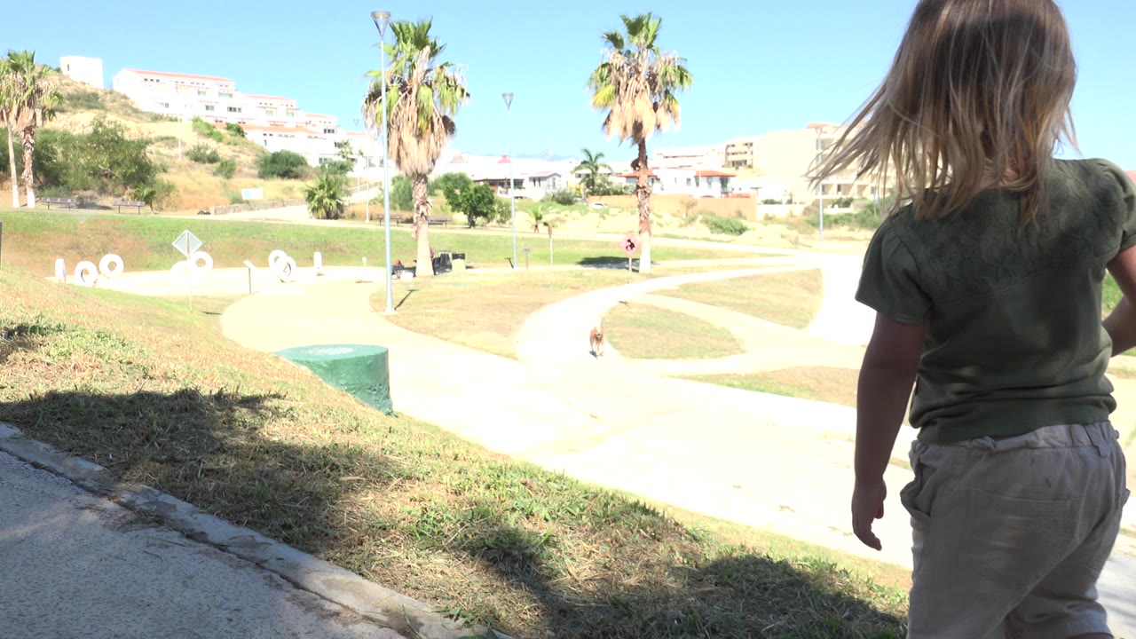 The child remains near the foreground shade while pale walkways, mowed grass, palm trees, and hillside buildings fill the Grand Junction park background.