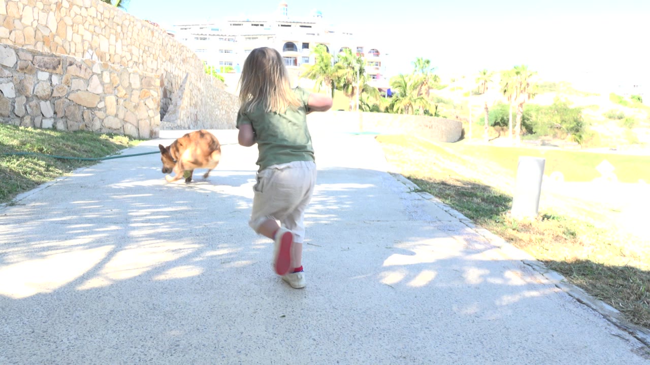 Young girl runs after a brown dog on a sunny concrete path.