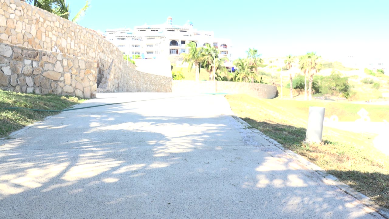 At 0:00 in Grand Junction, Colorado, the frame shows an empty sunlit paved path beside a stone wall, grass, bollards, palm-like trees, and distant buildings with no visible people.