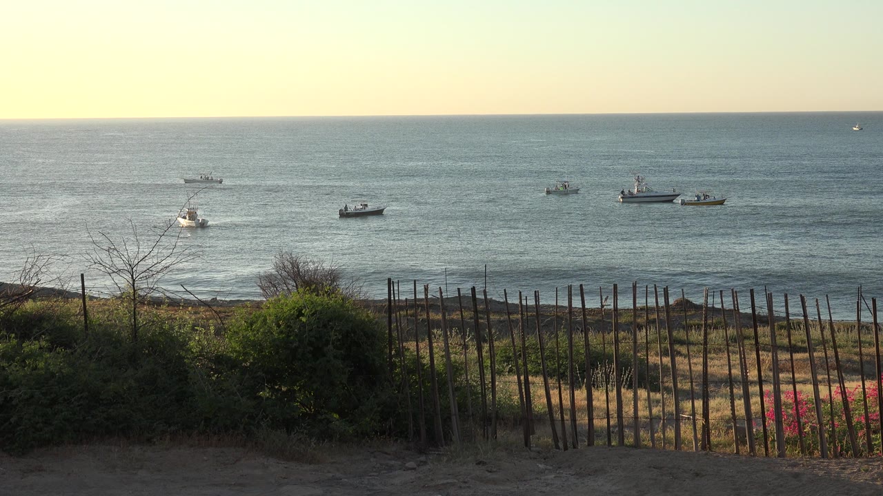 Boats offshore beyond a sandy dune and slatted fence.