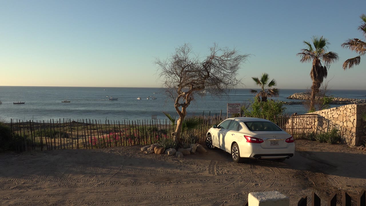 White sedan parked on dirt near water with boats offshore.