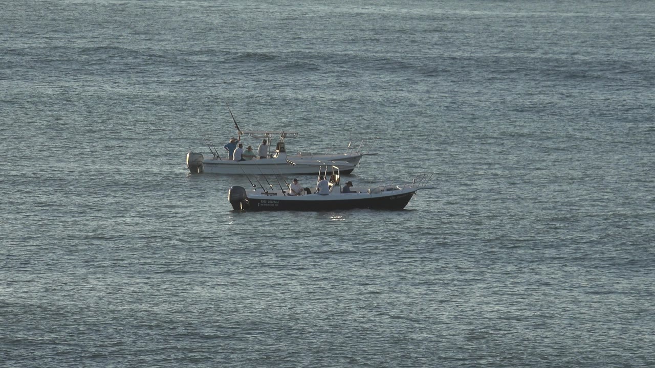The two boats remain near the upper center of the frame on blue-green rippled water, with distant occupants visible as small figures.