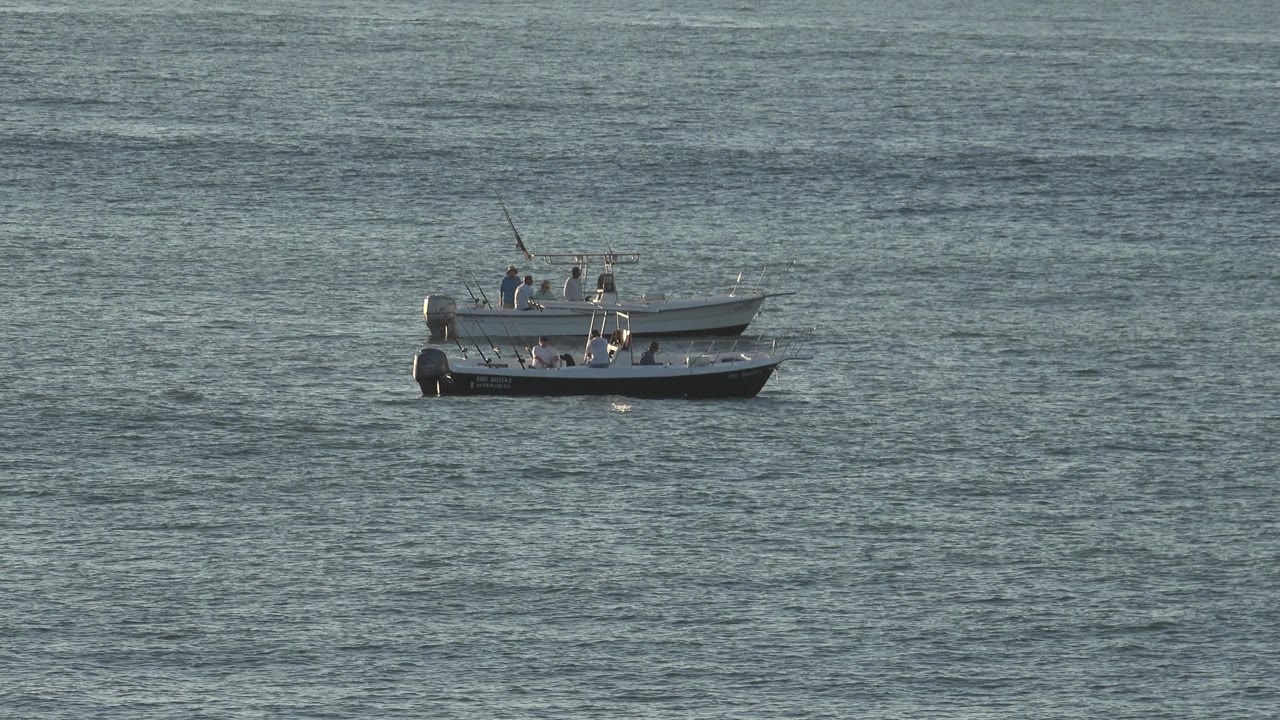 Two motorboats carrying visible people remain distant on gray-blue water, with fishing rods upright and light catching the white upper hulls.