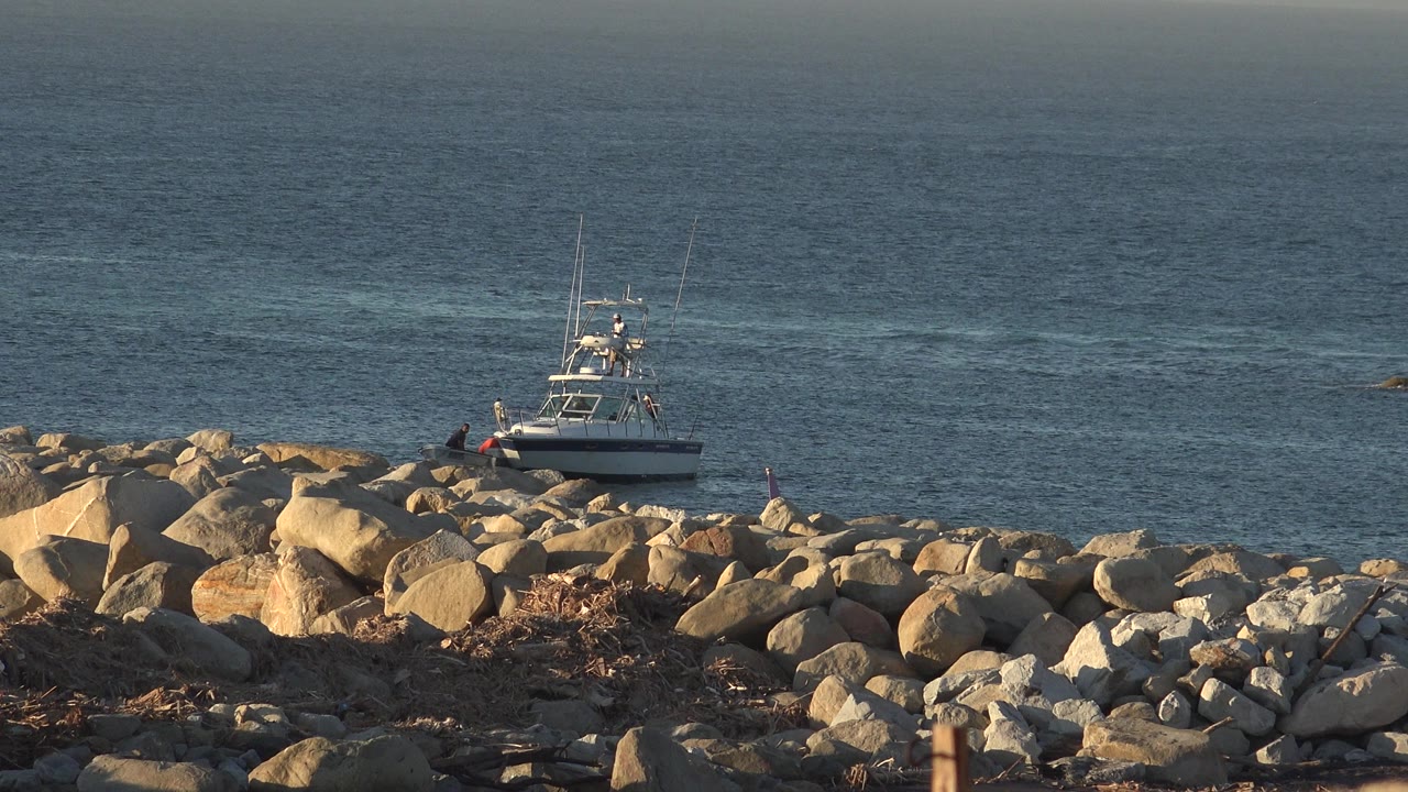 A small white boat sits off a rocky shoreline in Grand Junction, Colorado, with tan boulders in the foreground and open blue water behind it.