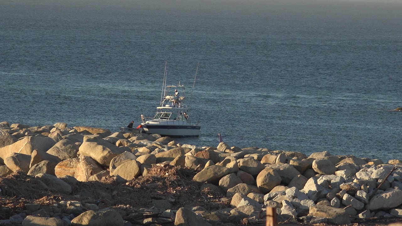 The boat remains beside the rock breakwater in Grand Junction, Colorado, while two distant people stand near the water and ripples spread across the bay.