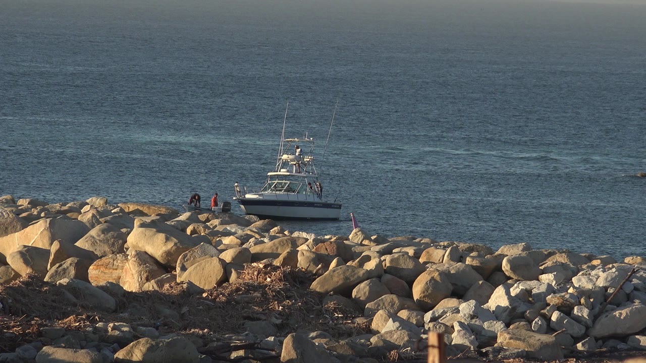 Small boat near a rocky shoreline with two people on the rocks.