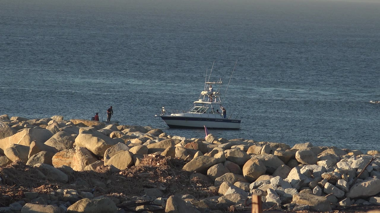 Two people near rocks with a white boat beyond them on open water.