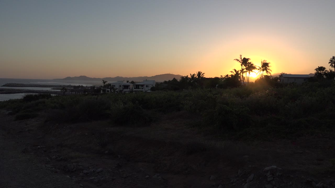 Sunset over water, hills, palms, brush, and distant houses.