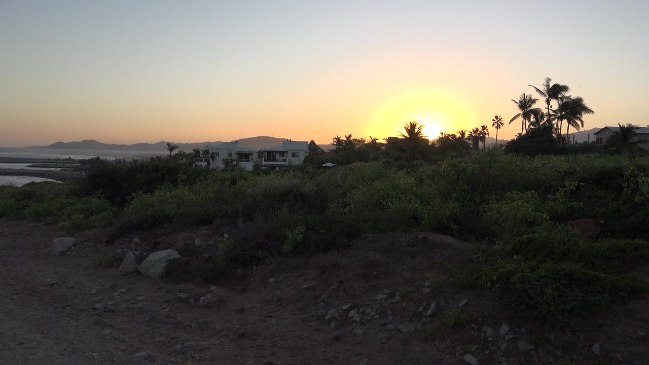 Sunset over shrubs, rocks, palm silhouettes, and a small house on a hillside.