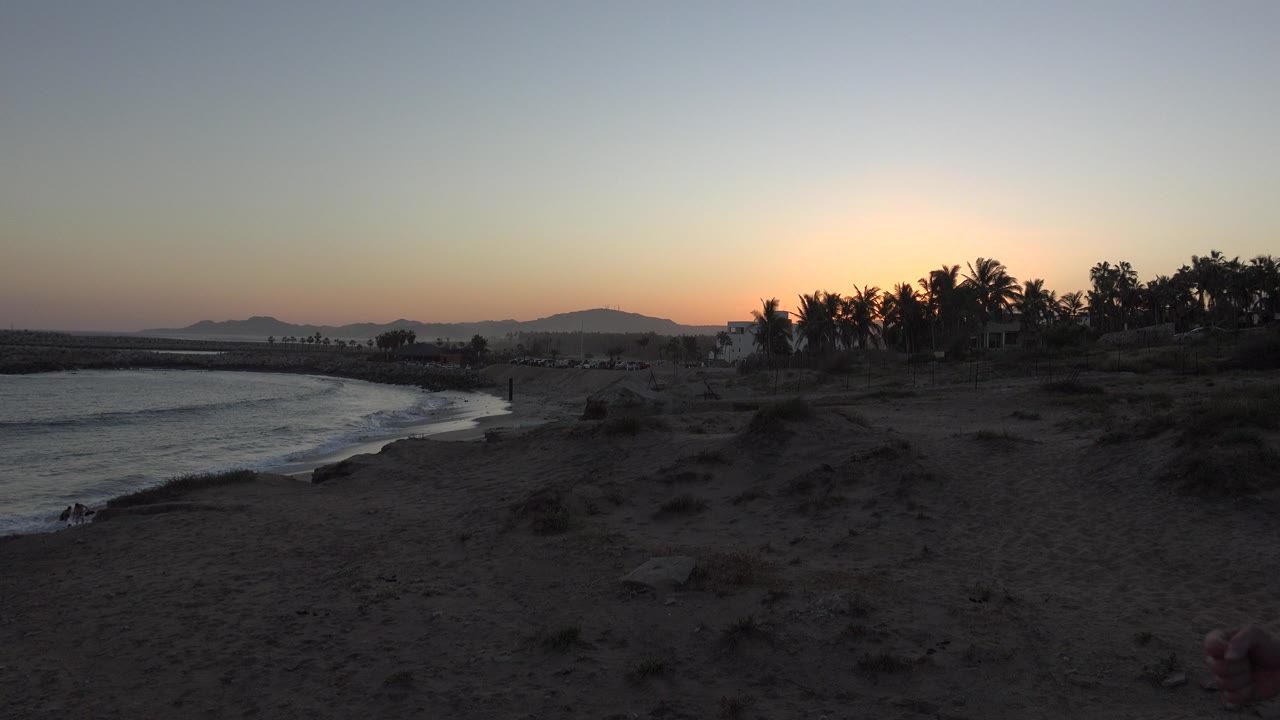 Second frame of the Grand Junction, Colorado beach scene, with the curving shoreline more centered, low waves on the left, dark sandy foreground, silhouetted palms on the right, and no visible people.