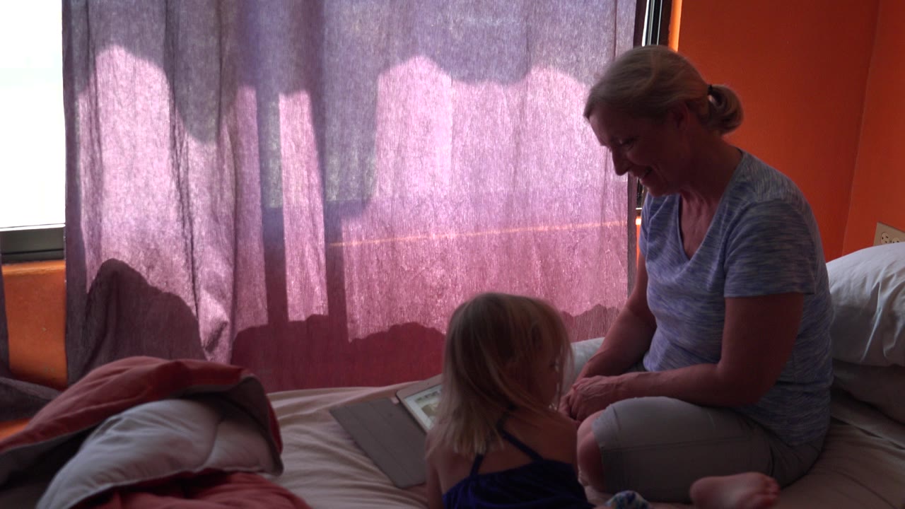 In Grand Junction, Colorado, a mother sits on a bed facing a young daughter near a tablet, with purple curtains glowing behind them.