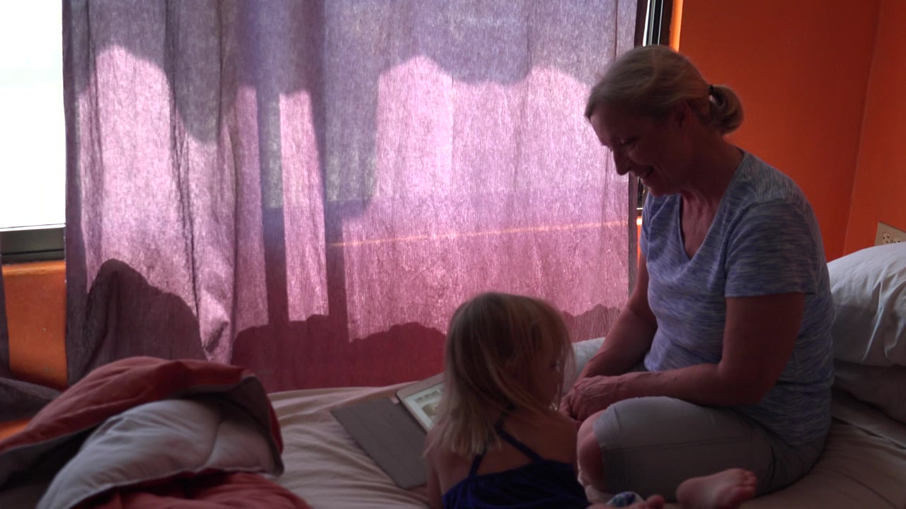 In Grand Junction, Colorado, the mother leans toward the child on the bed while the tablet rests nearby and the curtained window backlights the room.