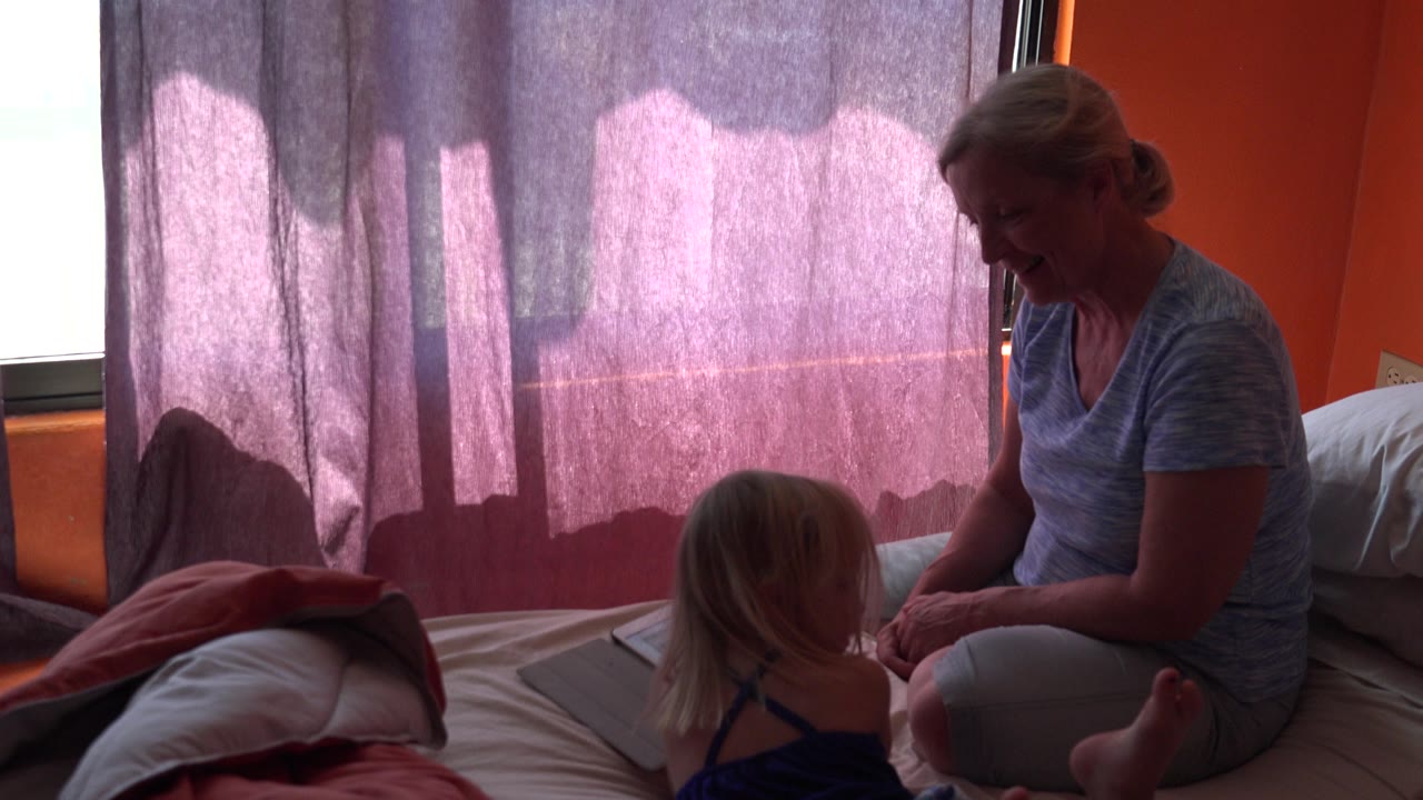 Child faces the seated mother beside a tablet on the bed.