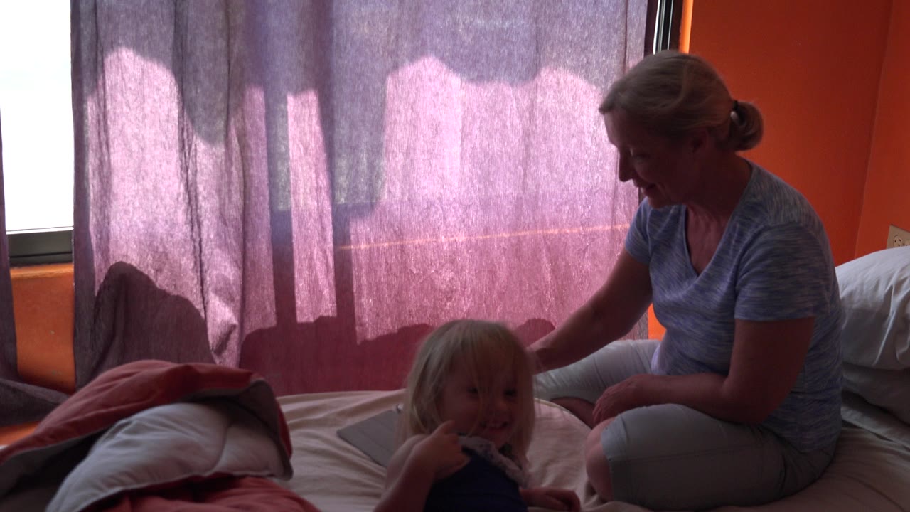 At 0:00 in Grand Junction, Colorado, a mother sits on a bed beside her young daughter near purple curtains and orange walls.