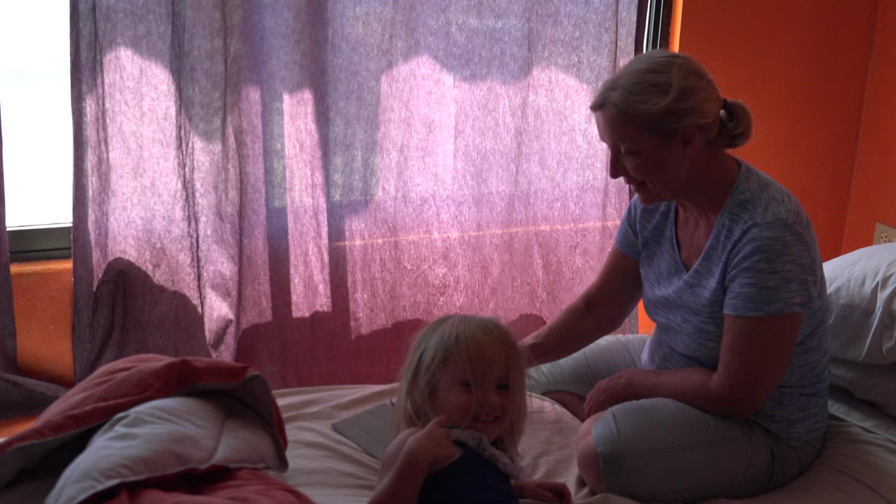 At 0:01, the young daughter faces toward the camera while her mother remains seated on the bed in the orange room in Grand Junction, Colorado.