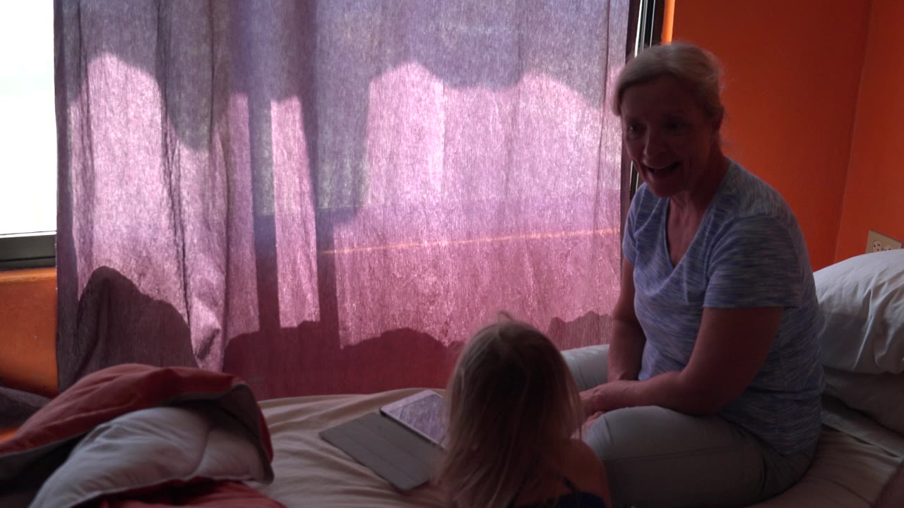 In Grand Junction, Colorado, an older woman sits on a bed beside a young girl, with a bright curtained window and orange wall behind them.