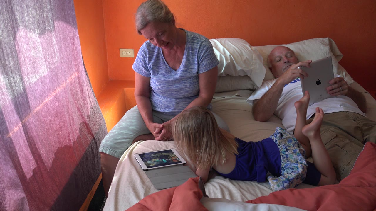 The child remains stretched across the bed in the foreground as the seated woman looks down and the reclining man studies a tablet in the Grand Junction home interior.