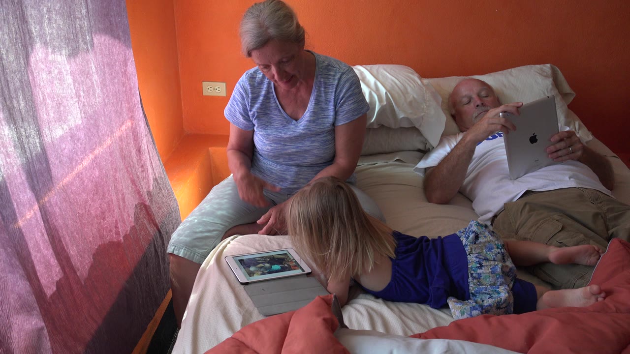 In Grand Junction, Colorado, the child remains stretched across the bed watching a tablet while the older man holds an Apple tablet and the older woman looks downward.