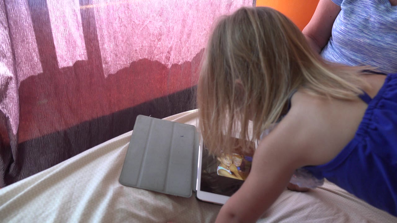 Blond child leaning over a tablet beside an adult indoors.