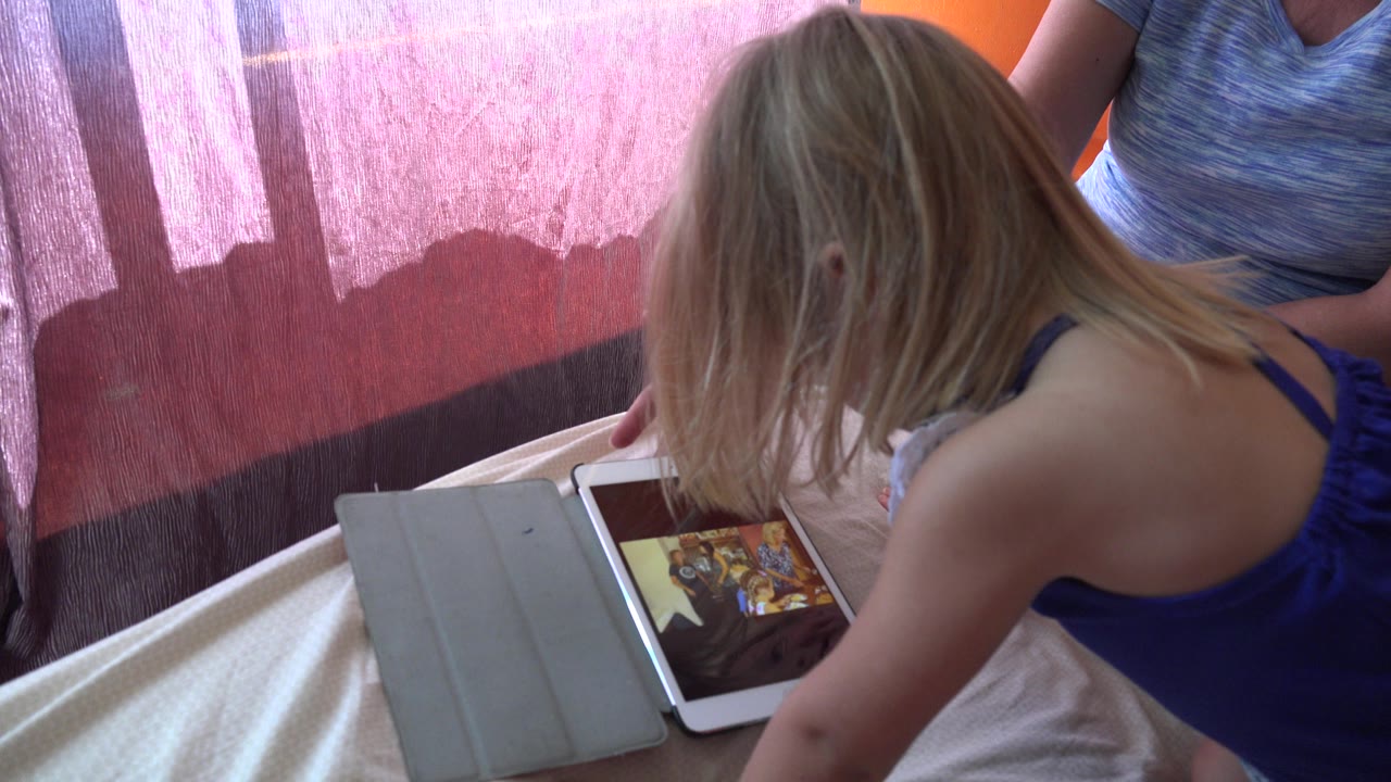 At 0:00 in Grand Junction, Colorado, a blonde little girl in a blue top leans over a white tablet on a bed while an adult woman is partly visible near pink curtains.