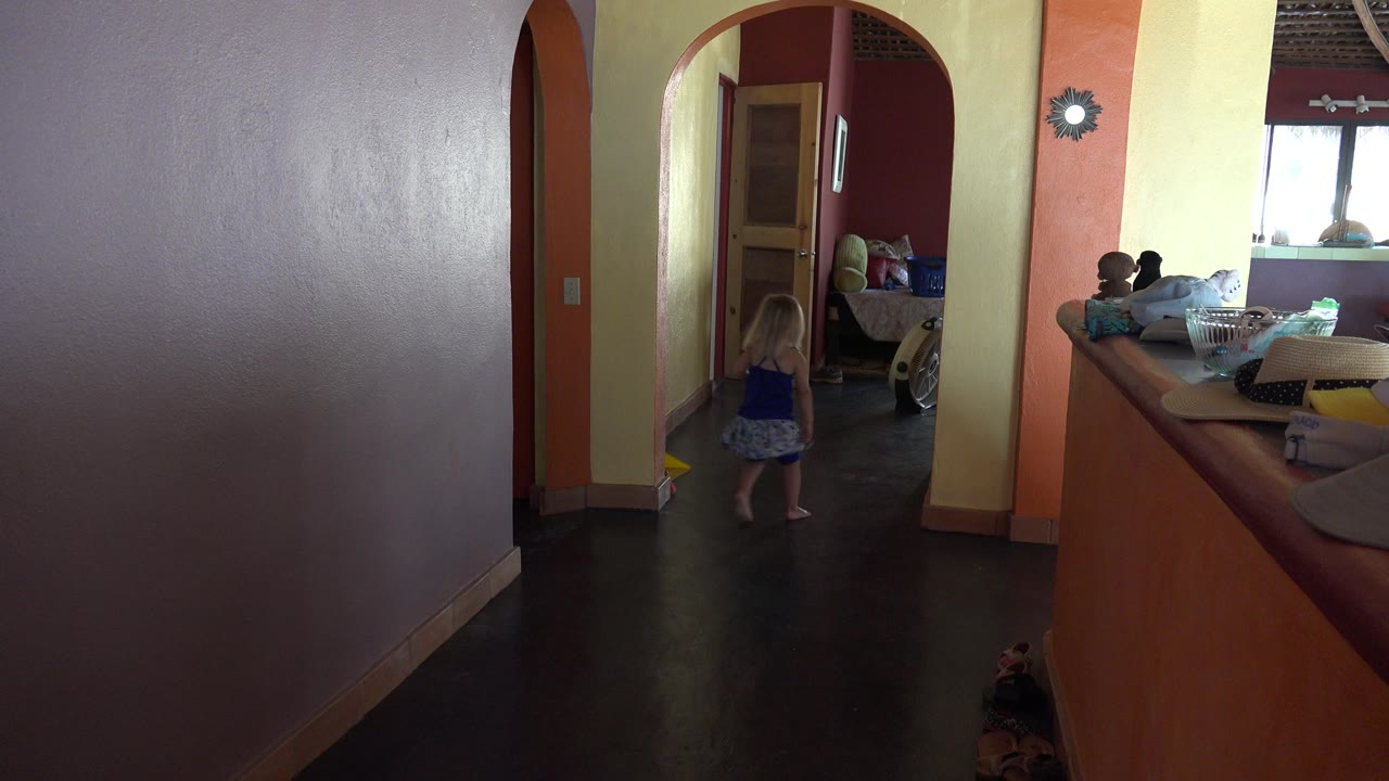 The young girl moves deeper through the arched doorway in the Grand Junction interior while a seated person remains near the couch or daybed by the window.