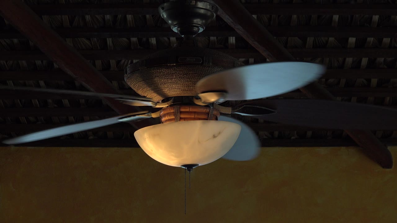 The fan blades blur as they spin around a warm ceiling light beneath the dark roof structure, with no visible people.
